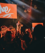 A performer stands on stage with bright red lighting and screens behind them, engaging with a lively audience. The crowd is raising their hands and capturing the moment on their phones. Disco balls and stage lights enhance the energetic atmosphere.